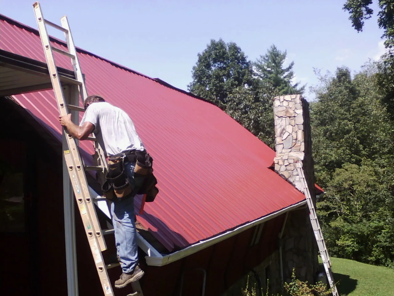 Roofer on a ladder beside a red metal roof and stone chimney — Soffit & Fascia Repair by Roofing Brothers in Linton